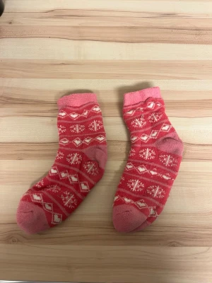 Rosa mönstrade sockor med snöflingor - Ett par rosa sockor med vitt vintermönster och snöflingor. Sockorna har förstärkta tår och hälar i en mörkare rosa nyans. Perfekta för att hålla fötterna varma under kyliga dagar.