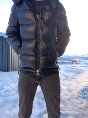 Svart glossy pufferjacka från Polo Ralph Lauren - Svart pufferjacka från Polo Ralph Lauren med röd logga på bröstet. Jackan har huva, dragkedja framtill och två sidofickor med dragkedja. Den är quiltad och har en glansig finish som ger en cool look. Perfekt för kalla dagar.