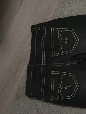 Svarta bootcut jeans med kontrastsömmar - Snygga svarta bootcut jeans med vita kontrastsömmar och unika broderade bakfickor. Jeansen har klassisk femficksdesign och bälteshällor. Materialet är troligtvis bomullsdenim med stretch för extra komfort.