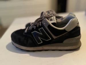 Svarta New Balance 574 sneakers - Klassiska New Balance 574 sneakers i svart mocka och mesh med grå detaljer och vita skosnören. Ikoniska N-loggan på sidan och bekväm vadderad häl. Snygg retrostil som passar till det mesta.