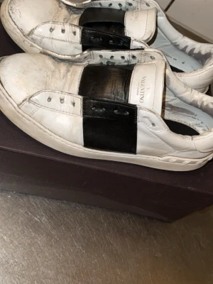 Valentino vita sneakers med svart detalj - Säljer ett par vita sneakers från Valentino med en bred svart läderdetalj över mitten. Klassisk låg modell med vita snören och diskret logga på plösen. Snygg och stilren design i läder som passar till många outfits.