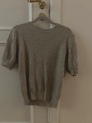 Beige stickad tröja med puffärm - Beige stickad tröja med korta puffärmar och rund halsringning. Tröjan har grovt garn som ger en avslappnad look. Perfekt för lager-på-lager och enkel att matcha med jeans eller kjol.