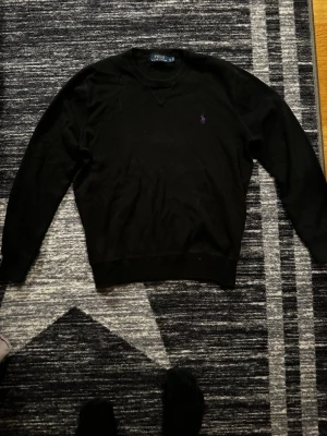 Svart stickad tröja från Polo Ralph Lauren - Svart stickad tröja från Polo Ralph Lauren med lila broderad logga på bröstet. Klassisk rund halsringning och ribbade muddar vid ärmslut och nederkant. Perfekt för lager-på-lager och stilrena outfits.