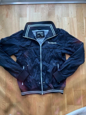 Marinblå vindjacka från Jack & Jones - Snygg marinblå vindjacka från Jack & Jones med vit dragkedja och vita detaljer på krage och muddar. Jackan har hög krage, två fickor med tryckknappar och ribbade muddar. Tillverkad i lätt, vindtätt material som passar perfekt till blåsiga dagar.