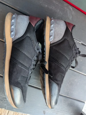 Svarta sneakers från Valentino Garavani - Snygga svarta sneakers från Valentino Garavani med detaljer i mocka, mesh och läder. Skorna har grå och vinröda partier vid hälen samt coola nitar på sulan. Klassisk snörning och gummisula för skön komfort.