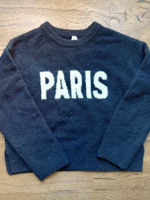 Stickad tröja med PARIS-tryck - Mysig svart stickad tröja. Tröjan har rund halsringning, långa ärmar och ribbade muddar vid ärmslut och nederkant. 🖤