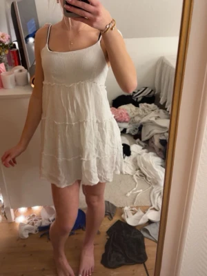 Vit kort sommarklänning med smala band - Söt vit kort klänning med smala axelband och lätt volang i kjolen. Klänningen har en luftig passform och är perfekt för varma dagar. Materialet känns mjukt och lätt, troligen bomull eller linne. Enkel och stilren design som passar till många tillfällen.