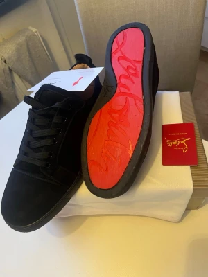 Svarta sneakers från Christian Louboutin - Snygga svarta sneakers från Christian Louboutin med klassisk röd sula och diskret logga. Skorna har en låg profil, svarta skosnören och är tillverkade i mocka. Perfekta för dig som vill ha något exklusivt och stilrent med en ikonisk touch.