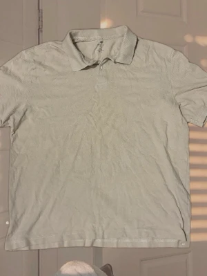 Vit pikétröja med krage - En stilren vit pikétröja med klassisk krage och korta ärmar. Tröjan har knappar vid halsen och är tillverkad i ett mjukt bomullsmaterial som känns bekvämt mot huden. Perfekt för en avslappnad men ändå snygg look.