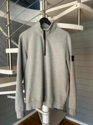 Grå halvzip Stone Island - Grå halvzip med dragkedja och hög krage från Stone Island. Tröjan har lång ärm och klassisk patch på vänster ärm. Tillverkad i mjukt bomullsmaterial, perfekt för en avslappnad och stilren look. Storlek L. Normal i passformen. 