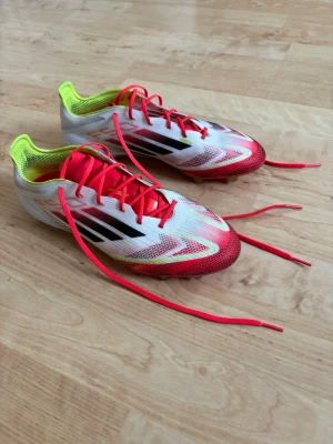 Adidas vita och röda fotbollsskor - Snygga Adidas fotbollsskor med vit bas, röda och neongula detaljer samt svarta ränder på sidorna. Skorna har röda snören och en lätt, ventilerande ovandel i syntetmaterial. Perfekta för snabba vändningar på planen.