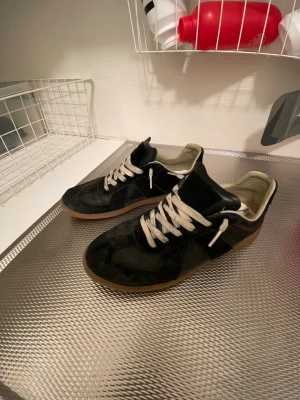 Svarta Maison Margiela Gats  - Säljer mina svarta Gats med vit/beigea snören. Storlek 42. Mycket bra skick, inga som helst defekter på själva skon, endast lite smutsiga, men går ju lätt att tvätta bort. Priset är inte hugget i sten, hör av er!