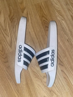 Vita Adidas tofflor med svarta ränder - Säljer ett par klassiska vita Adidas tofflor med tre svarta ränder och Adidas-logga på sidan. Tillverkade i syntetmaterial med bekväm fotbädd och öppen tå. Perfekta för stranden eller att chilla hemma. Storlek 39