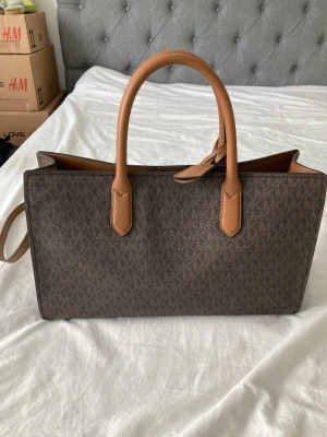 Brun Michael Kors handväska plus plånbok  - Snygg handväska från Michael Kors i brunt konstläder med MK-mönster och ljusbruna handtag. Väskan har guldfärgade detaljer och ett hänglås med MK-logga. Rektangulär form och både handtag och axelrem för olika stilar. Ingår plånbok.