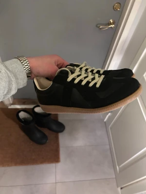 Maison Margiela gats  - Riktig bra skick endast använda ett par gånger och säljer för att dom var lite för små för mig. Snygga svarta sneakers med beige sula och vita snören. Klassisk låg modell med retrovibbar och stilren design. Ovandelen är i mocka och läder, vilket ger en schysst kontrast och premiumkänsla. Perfekta för dig som gillar minimalistisk stil. Har ingen låda kvar