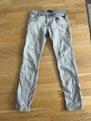 Ljusgrå slitna jeans från Replay - Säljer ett par ljusgrå jeans från Replay med skinny fit och slitningar på båda knäna. Jeansen har fem fickor, dragkedja och knapp i midjan samt tydliga kontrastsömmar. Perfekta för dig som gillar en avslappnad och trendig stil.