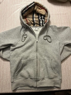 Grå zip-hoodie från Burberry - Snygg grå zip-hoodie från Burberry med klassiskt rutigt foder i huvan. Hoodien har dragkedja framtill, två fickor och justerbara snören i huvan. Tillverkad i mjuk bomullsblandning för extra komfort. Perfekt för en avslappnad och trendig look.