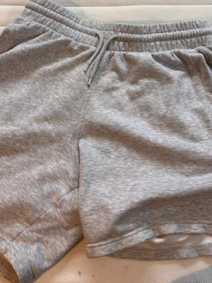 Grå mjukisshorts från H&M - Säljer ett par grå mjukisshorts från H&M med snörning i midjan och avslappnad passform. Shortsen har elastisk midja och är tillverkade i en mjuk bomullsblandning, perfekta för chill dagar eller träning.