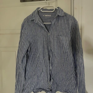 Blåvit randig skjorta från Zara - Snygg blå skjorta från Zara med vita vertikala ränder. Skjortan har klassisk krage, knäppning framtill och en bröstficka. Tillverkad i ett lätt och luftigt material som passar perfekt till varmare dagar. 