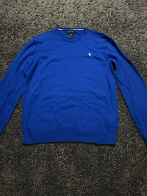Blå tröja från Polo Ralph Lauren - Klassisk blå tröja från Polo Ralph Lauren med rund halsringning och diskret broderad logga på bröstet. Tröjan har långa ärmar och ribbade muddar vid ärmslut och nederkant. Perfekt för en stilren och enkel look.