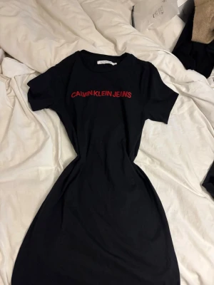 Svart t-shirtklänning Calvin Klein Jeans - Svart t-shirtklänning från Calvin Klein Jeans med röd logotyp på bröstet. Klänningen har korta ärmar och rund halsringning. Perfekt för en avslappnad och cool look. Materialet är mjuk bomull som känns skönt mot huden. (Pris går att diskutera)