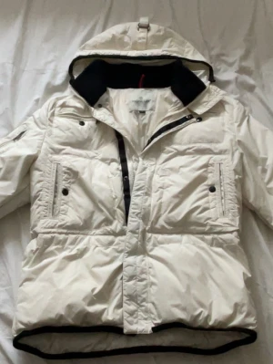 Vit pufferjacka från Moncler - Säljer en snygg vit pufferjacka från Moncler med huva och svart innerkrage. Jackan har två stora bröstfickor med knappar, dragkedja och tryckknappar framtill samt Moncler-logga på ärmen. Perfekt för kalla vinterdagar och riktigt stilren.
