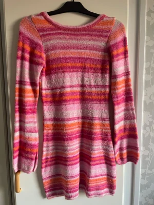 Randig stickad miniklänning i rosa och orange - Säljer en långärmad stickad miniklänning med färgglada ränder i rosa, orange, vitt och lila. Tröjan har en rund halsringning och är mjuk och stretchig i materialet. Den är öppen i ryggen. Perfekt för dig som gillar färg och vill sticka ut lite extra. Står att det är xxs men skulle säga att den är mer som en s. 