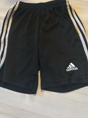 Svarta Adidas shorts med vita ränder - Svarta sportiga shorts från Adidas med klassiska vita ränder längs sidorna och Adidas-logga framtill. Perfekta för träning eller chill. Elastisk midja för skön passform.