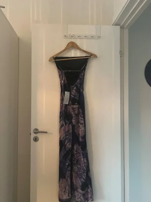 Lång mönstrad klänning med smala band - Säljer en lång klänning med smala axelband och ett abstrakt mönster i lila, svart och beige toner. Klänningen har en figurnära passform och är tillverkad i ett mjukt, stretchigt material som känns bekvämt mot huden. Perfekt för dig som vill sticka ut med en unik look.