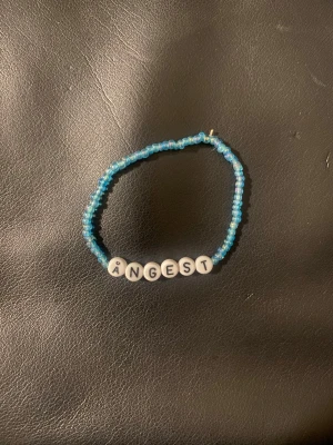 Blått pärlarmband med text - Ett handgjort armband med små blå pärlor och vita bokstavspärlor som stavar 'ÅNGEST'. Armbandet har en enkel och personlig stil, perfekt för dig som gillar unika accessoarer. Materialet är plastpärlor i olika nyanser av blått och vitt.