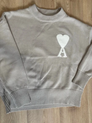 Beige knitted sweater from Ami - Snygge beige stickad tröja från Ami Alexandre Mattiussi med vitt hjärta och A-logga på bröstet. Tröjan har ribbade muddar vid ärmslut, hals och nederkant samt en avslappnad passform. Perfekt för dig som gillar stilrena och trendiga plagg.