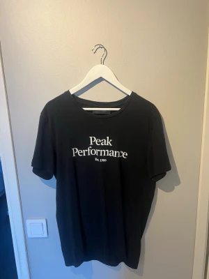 Svart Peak Performance t-shirt - Svart t-shirt från Peak Performance med vit logga tryckt på bröstet. Klassisk rund halsringning och korta ärmar. Perfekt för en avslappnad och stilren look.