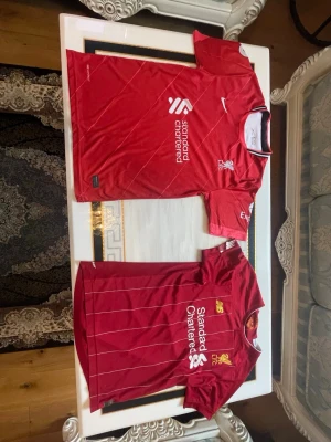 2 styckna Liverpool matchtröjor herr röd - Två röda Liverpool FC matchtröjor med vita detaljer och klubbmärke på bröstet. Ena tröjan har tryck med M. Salah och nummer 11 på ryggen. Båda har Standard Chartered som sponsor och är tillverkade i lätt, ventilerande material.