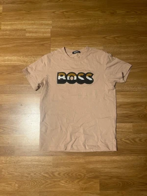 Beige BOSS t-shirt med logga - Snygg beige t-shirt från BOSS med stor svart och gul logga på bröstet. Klassisk passform med rund hals och korta ärmar. Perfekt till jeans eller shorts för en clean och enkel stil. Tillverkad i mjuk bomull för skön känsla hela dagen.
