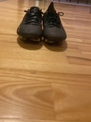 Svarta fotbollsskor med snörning - Säljer ett par svarta fotbollsskor med snörning och låg profil. Skorna har rund tå och platt sula med dobbar, perfekta för spel på gräsplan. Materialet är syntet och designen är stilren utan extra mönster.