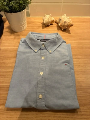Ljusblå skjorta från Tommy Hilfiger - Klassisk ljusblå skjorta från Tommy Hilfiger med button-down krage och vita knappar. Skjortan har lång ärm och den ikoniska lilla loggan broderad på bröstet. Perfekt till jeans eller chinos för en clean look.