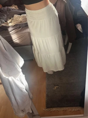 Vit långkjol med volanger - Säljer en vit långkjol med bred resår i midjan och flera volanglager. Kjolen har en luftig och flowy känsla, perfekt för varma dagar. Passar till crop tops och sneakers för en chill look.