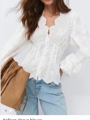 Vit blus med broderade blommor - Snygg vit blus med broderade blommor och hålmönster. Blusen har en figurnära passform, knappar framtill och vågig kant längs nederkanten och halsen. Långärmad med ballongärmar och tillverkad i ett lätt bomullstyg.