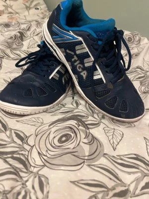 Pingis skor från stiga  - Säljer ett par blåa inomhusskor från Stiga med vita och silverfärgade detaljer. Skorna har snören, ventilerande meshpartier och vit sula som passar perfekt för sporter inomhus, speciellt pingis.  Insidan är fodrad med blått tyg för extra komfort. Litet hål på ena skon, vet inte hur det gick till? Skorna är slitna men dom har gjort ett bra jobb!OBS: Om ni vill köpa så kommer ni få en Yasaka låda med skorna i. Så att i vet! 