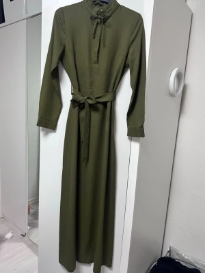 Olivgrön långklänning med knyt - Stilren olivgrön långklänning med lång ärm, knytband i halsen och avtagbart midjebälte. Klänningen har en rak passform och enkel design som ger en clean look. Perfekt för dig som gillar minimalistisk stil.