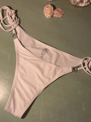 Vit Bikiniunderdel - Snygg vit bikiniunderdel med trendiga knytband i sidorna och små hjärtformade metalldetaljer. Tillverkad i stretchigt material som sitter bekvämt. Perfekt för strandhäng eller poolparty. 