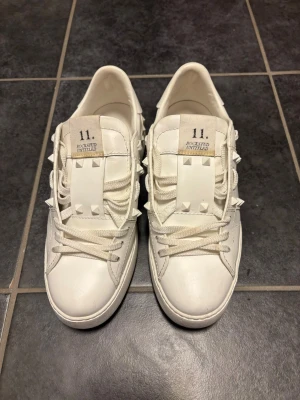 Vita Valentino Garavani sneakers med nitar - Säljer ett par vita Valentino Garavani sneakers med ikoniska pyramidformade nitar längs sidorna. Skorna har snörning, platt sula och är tillverkade i skinn. Perfekta för dig som gillar stilrena och exklusiva sneakers. Använd max 10 gånger, skickas i original låda med extra snören. Skriv ett meddelande om fler bilder önskas. Köpa på NK för 7000kr
