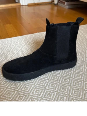Svarta Chelsea boots i mocka - Säljer ett par stilrena svarta Chelsea boots i mocka med elastiska paneler på sidorna och dragflikar både fram och bak. Skorna har en klassisk siluett och tjock sula som ger en modern touch. Perfekta för dig som gillar minimalistisk och clean stil.