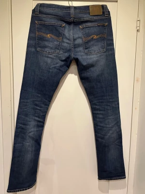 Mörkblå jeans från Nudie Jeans - Säljer ett par mörkblå jeans från Nudie Jeans med klassisk femficksmodell och snygga kontrastsömmar på bakfickorna. Jeansen har rak passform och är tillverkade i slitstark denim. Perfekta för en avslappnad och stilren look.