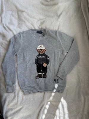 Grå stickad tröja med Polo Bear - Stickad tröja i ljusgrått från Polo Ralph Lauren med ikonisk Polo Bear-motiv på framsidan. Rund halsringning och ribbade muddar. RL-broderi vid nederkanten. Motivet har detaljer i svart, vitt och brunt. Bröst:45cm längd:56cm arm:56cm