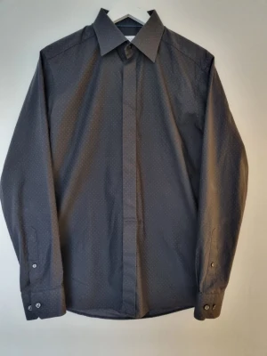 Eton Skjorta Stl 41 - Lätt mönstrad skjorta från Eton i färgen svart och med en kroppsnära passform. Den är i mycket bra skick och har inga tecken på användning. Kragomkretsen är 41 cm, vilket motsvarar ungefär en medium. Hör av dig om du har fler frågor så svarar jag gladeligen:)