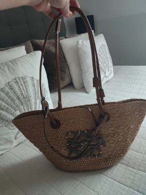 Stråväska från Loewe med läderdetaljer - Säljer en snygg stråväska från Loewe med bruna läderhandtag och dekorativt lädermärke framtill. Väskan har en korgformad design och är perfekt för dig som gillar en naturlig och trendig look. Passar till sommarens alla outfits!