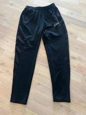 Svarta Nike Dri-FIT träningsbyxor - Säljer ett par svarta träningsbyxor från Nike med Dri-FIT-material som håller dig torr under träningen. Byxorna har resår i midjan och en vit Nike-logga på låret. Perfekta för sport eller gympass.