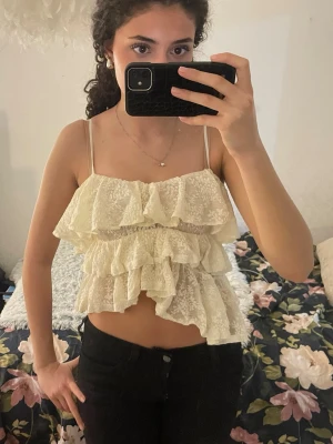Krämvit volang linnetopp i spets - Säljer en krämvit linnetopp med flera lager volanger och smala axelband. Toppen är croppad. Använd väldigt få gånger och i perfekt skick!💕