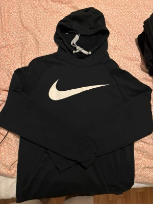 Svart Nike hoodie med vit logga - Säljer en svart hoodie från Nike med stor vit Swoosh-logga på bröstet. Hoodien har huva med vita dragsnören och en klassisk magficka framtill. Tillverkad i mjukt material som passar perfekt till en avslappnad stil.